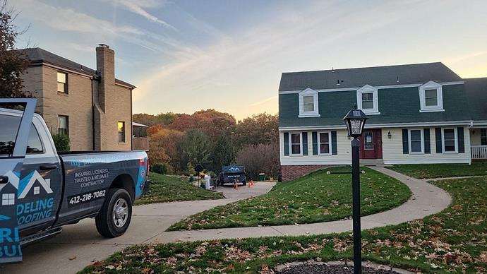 A professional roofing truck in the driveway of a client’s white house and black roof.